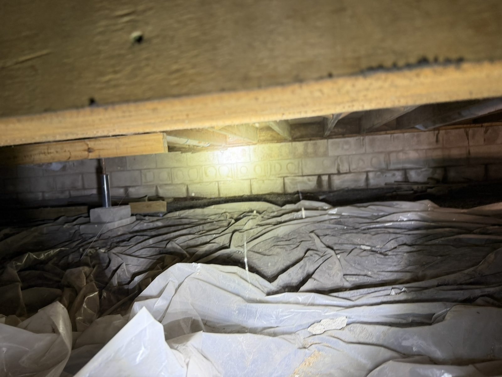 Dark crawl space before encapsulation showing sagging vapor barrier over a dirt floor and unsealed block foundation