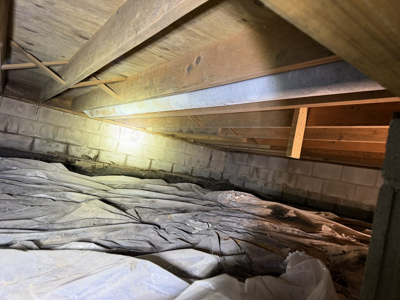 Crawl space before encapsulation with torn plastic vapor barrier, exposed dirt, and wood debris along a block foundation wall